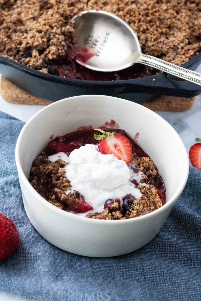 A perfectly sweet paleo berry cobbler made with three kinds of berries and #autumnsgold paleo granola in the topping. Add coconut whipped cream for the ultimate #paleo treat. #athousandcrumbs #paleo #berry #cobbler #strawberry #blueberry #raspberry #coconutwhippedcream